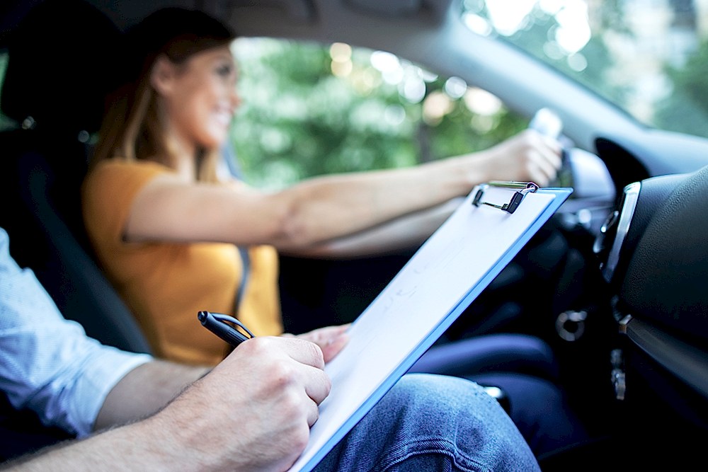 Close up view of driving instructor holding checklist while in background female student steering and driving car.